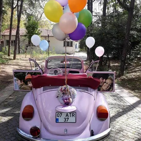Arkasında renkli balon demeti ve fotoğraflı tabelalar olan pembe cabrio Vosvos gelin arabası